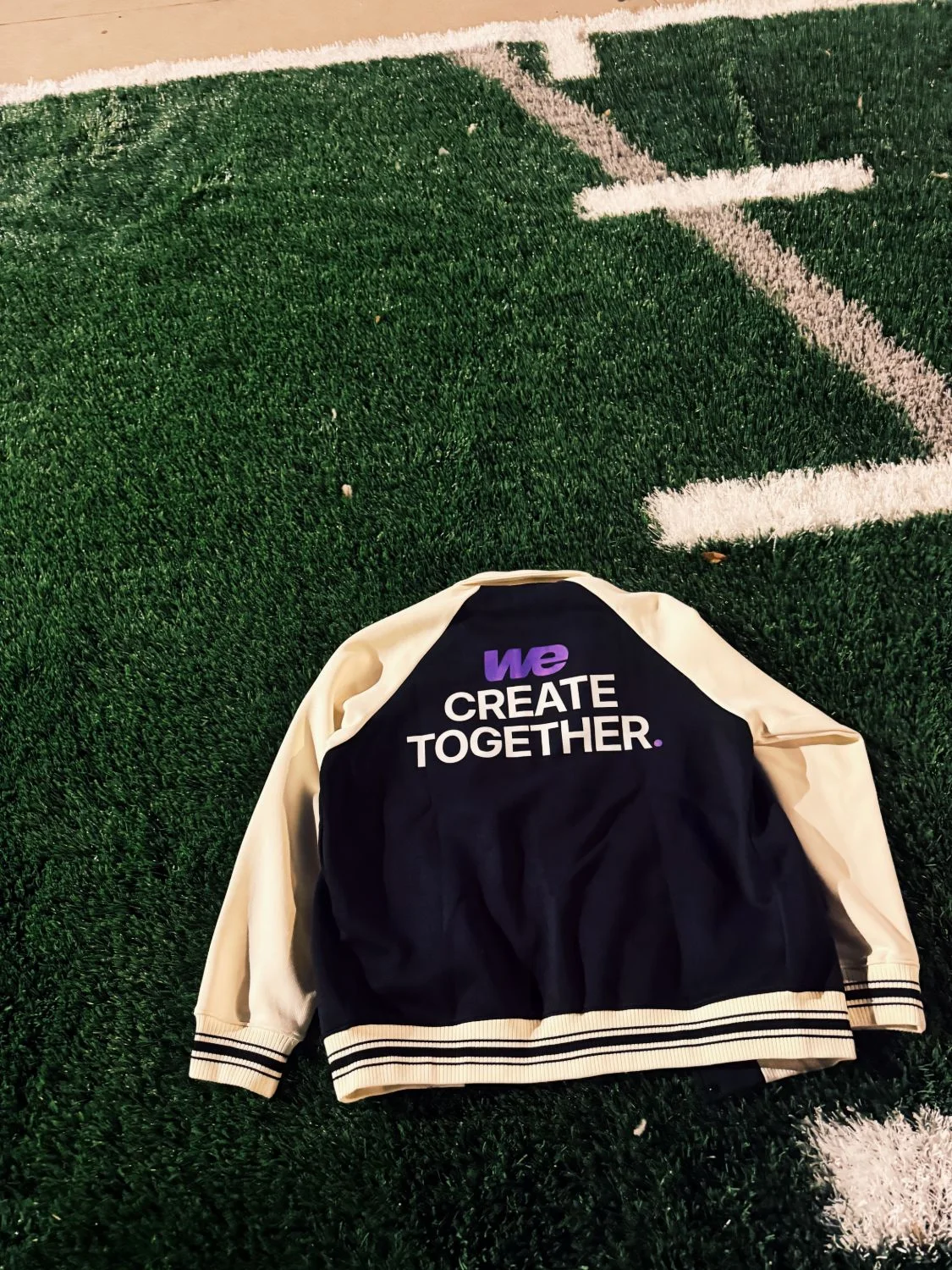 The Together Varsity lying on a football field at night: black body, cream sleeves, WE Create Together across the back