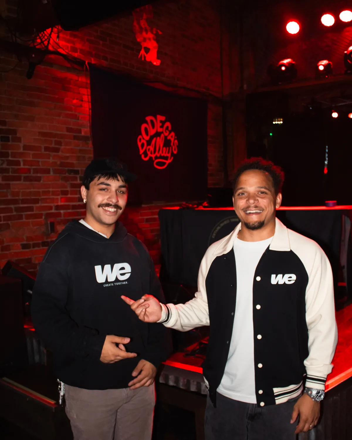 Two people at Bodega's Alley at night, one in the WE Hoodie and one in the Together Varsity